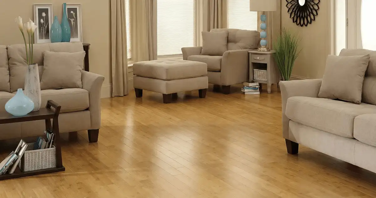 A living room with professionally installed bamboo flooring, beige sofas, a matching ottoman, a side table with a blue lamp, vases, framed pictures, and large windows with blinds and curtains in Anchorage, AK