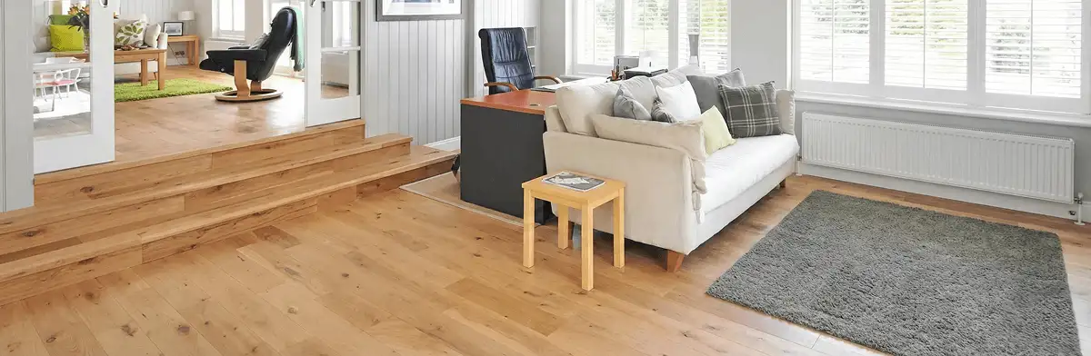 A modern living room with professionally installed laminate flooring, a beige sofa, a small side table, a gray rug, and large windows with white shutters in Anchorage, AK